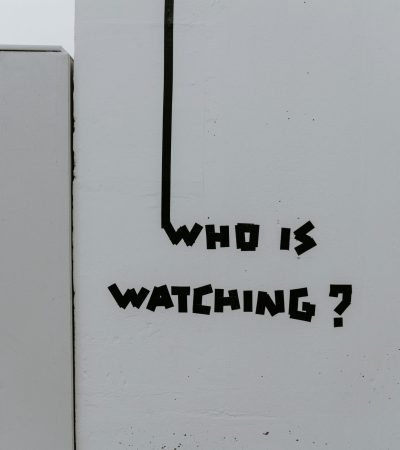 Instalação artística com a frase ‘Who is watching?’ feita em fita preta sobre parede branca; questiona vigilância e autenticidade