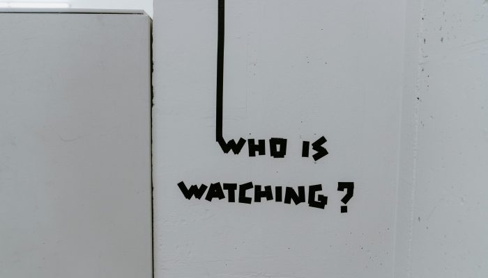 Instalação artística com a frase ‘Who is watching?’ feita em fita preta sobre parede branca; questiona vigilância e autenticidade
