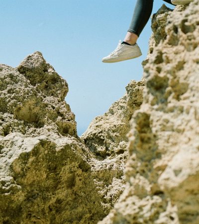 Pessoa sentada na borda de uma formação rochosa, com apenas parte das pernas e pés visíveis, usando tênis brancos e calça escura. O céu azul ao fundo destaca o contraste entre o corpo suspenso e as pedras irregulares.