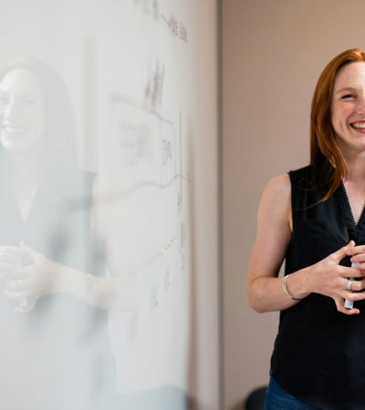 Mulher sorridente diante de um quadro branco, com postura profissional e confiante, simbolizando o equilíbrio entre acolhimento e responsabilidade no atendimento.