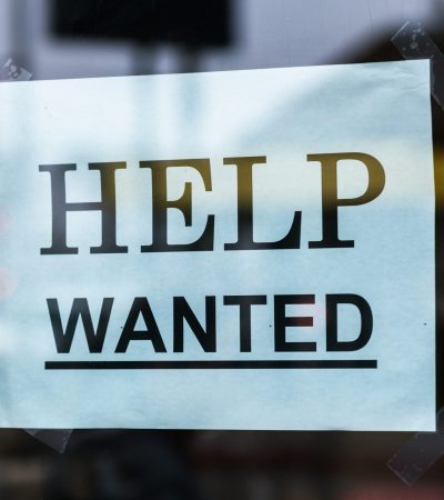 Cartaz com os dizeres 'HELP WANTED' em letras maiúsculas, colado com fita adesiva em uma vitrine de vidro. A placa indica que o local está contratando. O fundo mostra reflexos e luzes desfocadas de uma rua urbana.