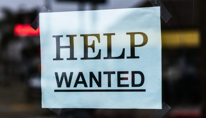 Cartaz com os dizeres 'HELP WANTED' em letras maiúsculas, colado com fita adesiva em uma vitrine de vidro. A placa indica que o local está contratando. O fundo mostra reflexos e luzes desfocadas de uma rua urbana.