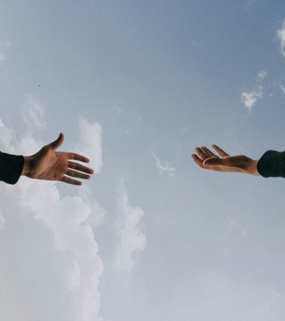 Duas mãos se estendem uma em direção à outra, contra um céu parcialmente nublado. Uma vem de cima, a outra de baixo. Ambas estão com mangas escuras, criando um contraste sutil com o azul do céu. A imagem evoca conexão, busca, apoio e o gesto simbólico de alguém sendo amparado — como na terapia.